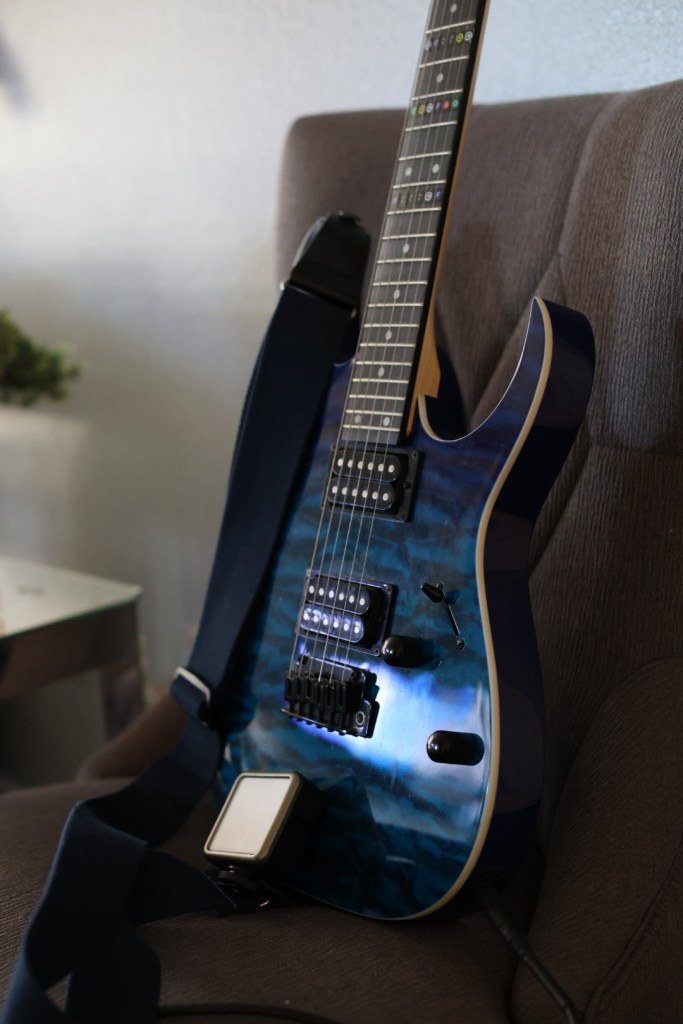 A blue electric guitar resting on a brown chair, with a guitar strap and a foot pedal nearby.